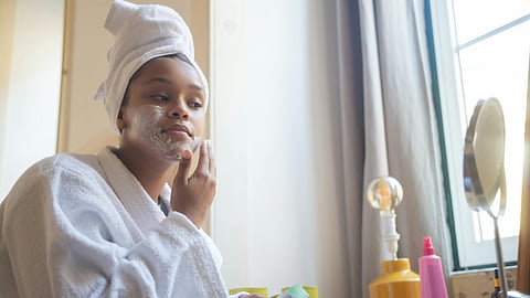 A Woman in White Bathrobe Applying a Cream on Her Face while Looking at the Mirror
